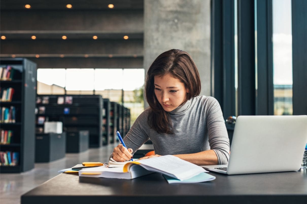 A woman sitting at a table, engrossed in her laptop and surrounded by books.